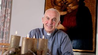 Ein Priester sitzt vor einem geschmückten Tisch mit Kerzen und einem religiösen Gemälde dahinter.