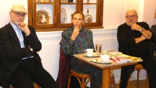 Eine Frau sitzt an einem Tisch mit zwei Tassen und Untertassen, einem Glas Wasser und Büchern. Hinter ihr steht ein hölzerner Schrank mit dekorativen Gegenständen. Ein weiterer Mensch sitzt links von ihr.