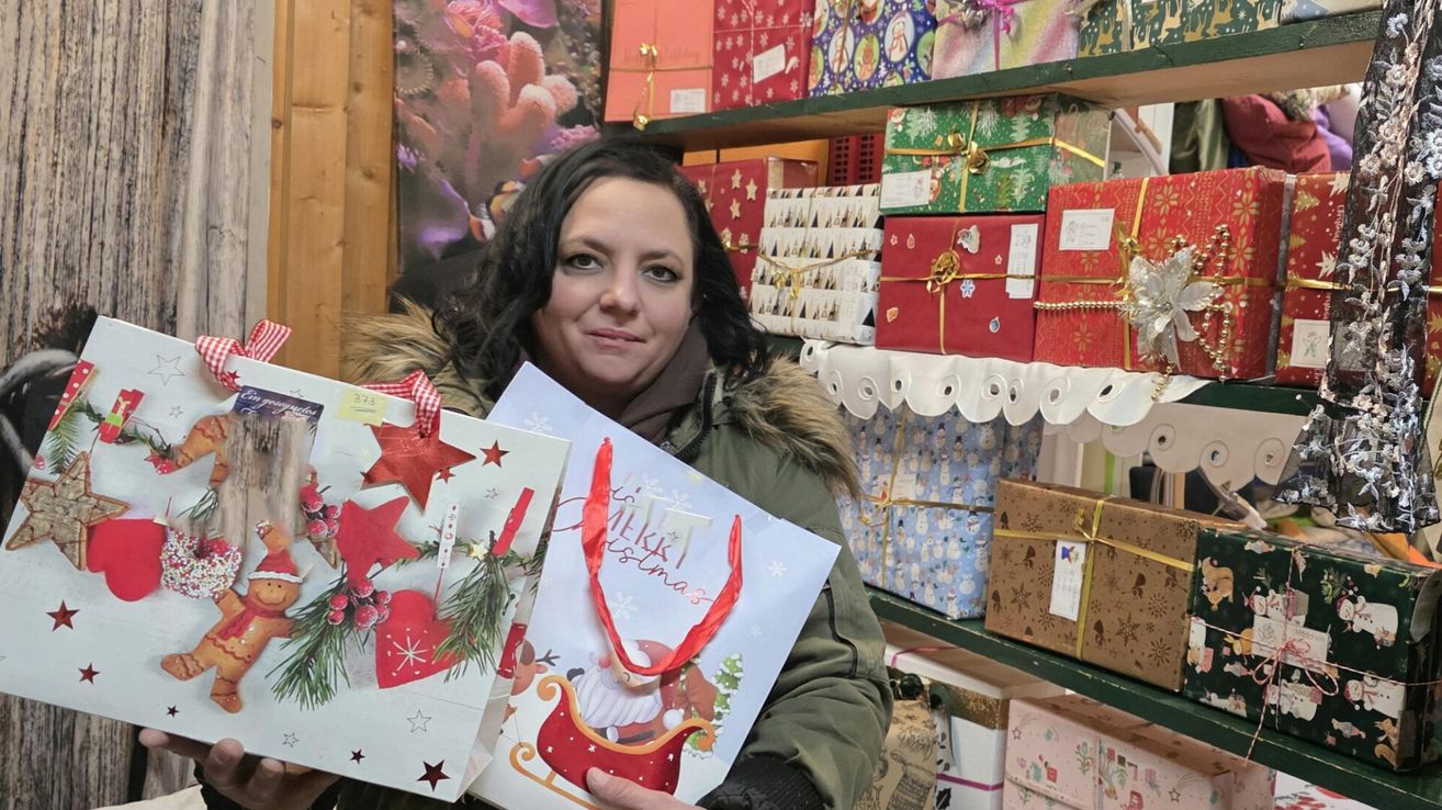 Eine Frau lächelt und hält zwei Weihnachtsgeschenktüten in einem Geschäft mit vielen verpackten Geschenken auf Regalen.