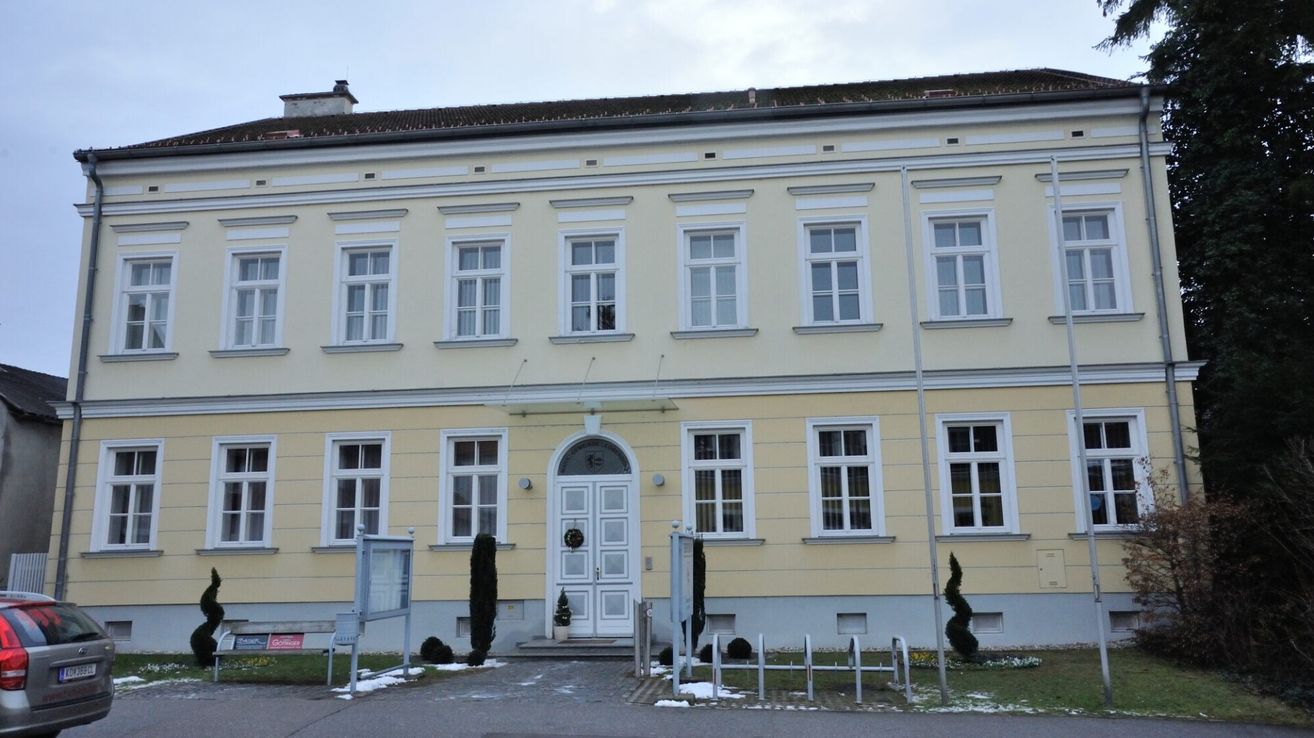 Ein großes, hellgelbes Gebäude mit mehreren Fenstern. Ein Kranz hängt an der Tür, und eine kleine Topfpflanze steht in der Nähe.