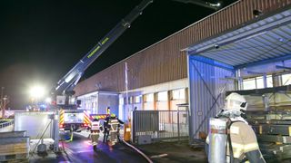 Feuerwehrleute reagieren auf einen Vorfall in einem Lagergebäude bei Nacht, mit einem Kran in der Nähe und mehreren Personen in Schutzausrüstung.