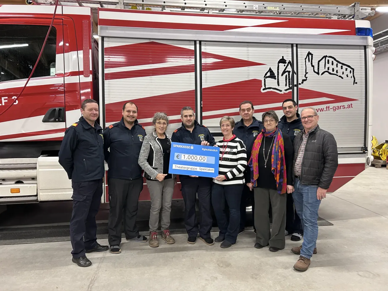 Eine Gruppe von Menschen in Uniformen steht vor einem Feuerwehrauto und hält einen blauen Spendenscheck über 1.000 Euro.