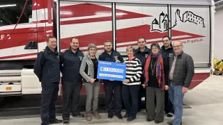 Eine Gruppe von Menschen in Uniformen steht vor einem Feuerwehrauto und hält einen blauen Spendenscheck über 1.000 Euro.