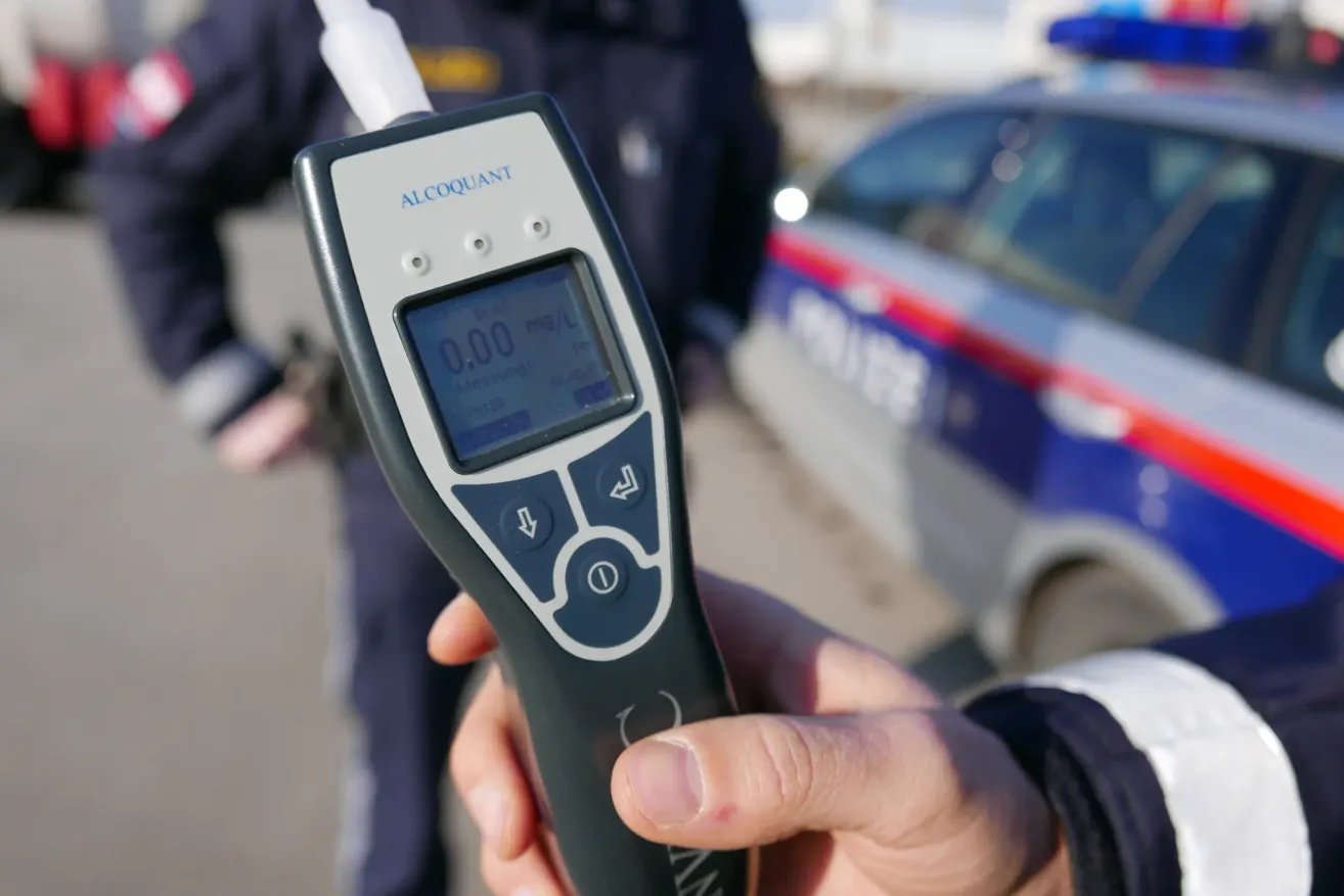 Eine Person hält ein Alcoquant-Gerät mit einer Anzeige von 0,00 mg/l und einem Polizeiauto im Hintergrund.