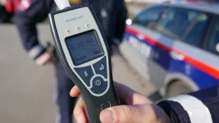 Eine Person hält ein Alcoquant-Gerät mit einer Anzeige von 0,00 mg/l und einem Polizeiauto im Hintergrund.