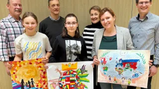 Eine Gruppe von Personen, darunter zwei Frauen und drei Mädchen, steht vor einer Holzwand, lächelt und hält bunte Zeichnungen hoch. Die Zeichnungen umfassen eine Weltkarte, eine Sonne und einen Globus. Die Frau rechts trägt eine Halskette und eine Brille.