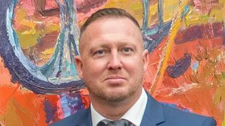 Ein Mann mit kurz geschnittenem Haar und Bart steht vor einem farbenfrohen Gemälde, er trägt einen blauen Anzug und eine Krawatte.