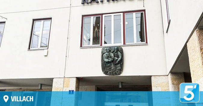 Ein weißes Gebäude mit roten Begrenzungen hat zwei Fenster und ein zentrales Fenster mit Blick auf ein Gebäude. Eine Plakette mit zwei Figuren ist an der Wand befestigt. Das Gebäude hat ein Namensschild mit der Aufschrift 'Villach'.