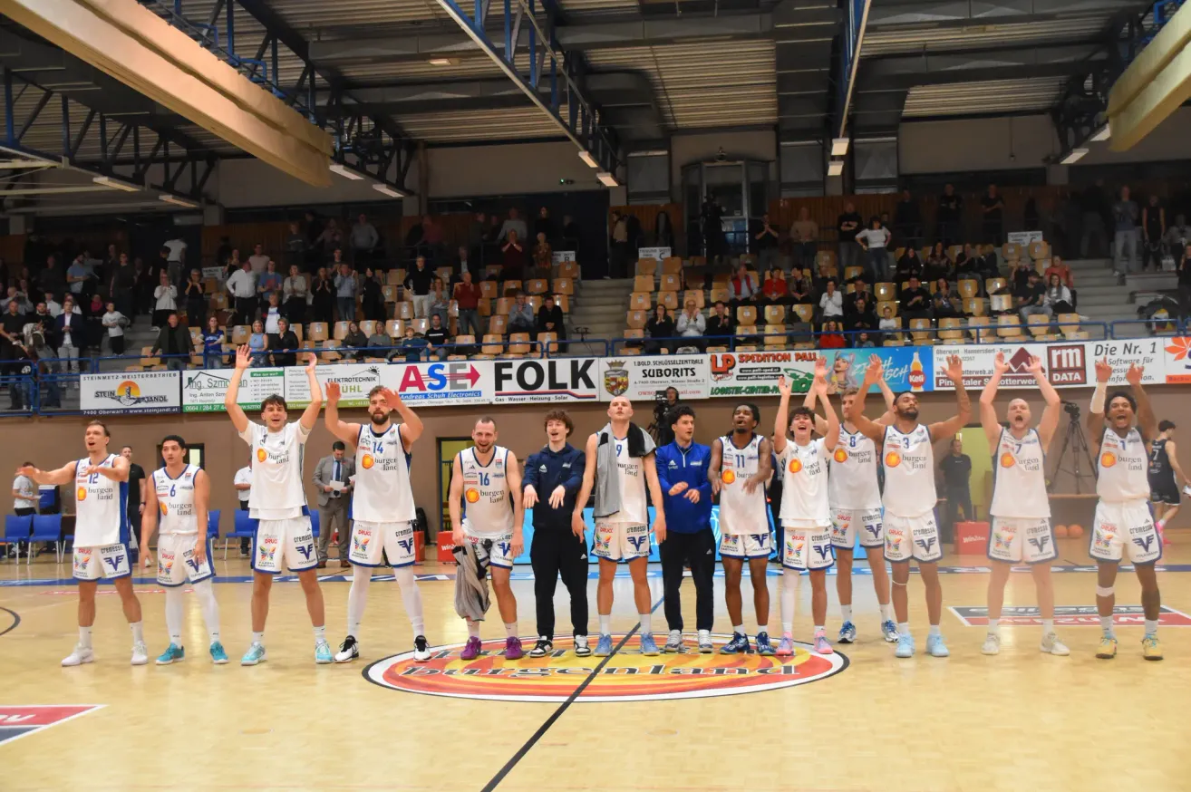 Basketballspieler feiern auf dem Spielfeld mit erhobenen Händen, umgeben von einem Publikum in einer Halle. Banner sind an den Seiten angebracht, und der Boden hat ein Logo.
