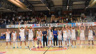 Basketballspieler feiern auf dem Spielfeld mit erhobenen Händen, umgeben von einem Publikum in einer Halle. Banner sind an den Seiten angebracht, und der Boden hat ein Logo.