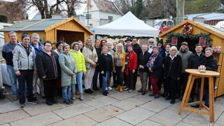 Eine Gruppe von Menschen, die warm gekleidet sind, posiert für ein Foto vor einem weißen Zelt und kleinen Holzhütten, wobei ein Gebäude und ein Auto im Hintergrund zu sehen sind.