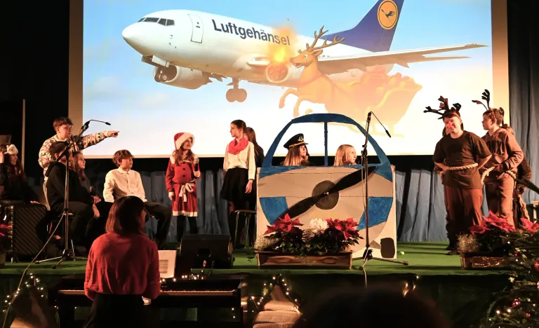 Kinder auf der Bühne, gekleidet in festliche Kleidung, führen vor einem großen Bildschirm vor, der ein Flugzeug und einen Renntier zeigt. Ein Klavier, Mikrofon und Blumenarrangement stehen auf der Bühne.