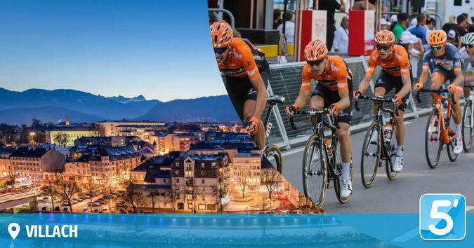 Eine Gruppe von Radfahrern in orangefarbenen Trikots fährt eine Stadtstraße hinunter. Im Hintergrund ist eine Stadt mit Bergen und einem Nachthimmel zu sehen. Der Text 'VILLACH' ist in der unteren linken Ecke angezeigt.
