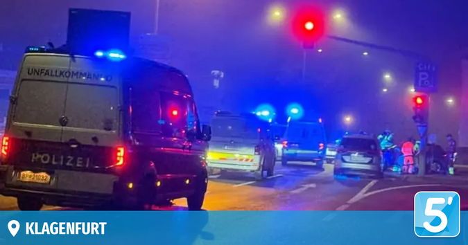 Einsatzfahrzeuge, darunter ein Polizeiwagen, sind auf einer nebligen Straße in der Nacht geparkt. Ampelanlagen und Schilder sind sichtbar, mit Fußgängern und anderen Fahrzeugen in der Nähe.