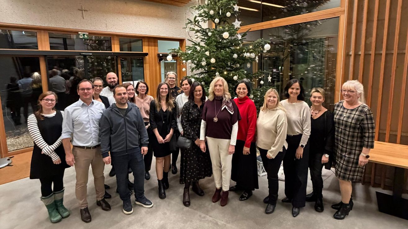 Eine Gruppe von Menschen posiert für ein Bild vor einem geschmückten Weihnachtsbaum. Sie lächeln und stehen vor einem Gebäude.