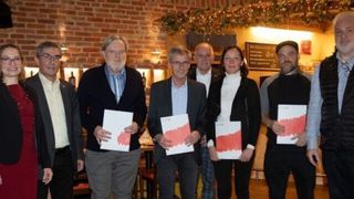 Fünf Personen stehen in einem Raum mit Ziegelwänden und halten Papiere. Ein Mann links hat einen weißen Bart und ein anderer in der Mitte trägt eine Brille. Zwei Frauen und ein Mann rechts schauen nach vorne.