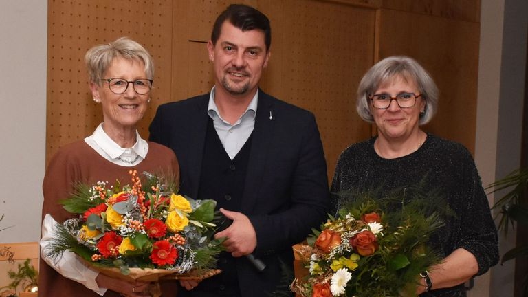Drei Menschen stehen lächelnd zusammen und halten Blumensträuße. Der Mann trägt einen dunklen Anzug, die Frauen tragen Brillen.