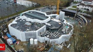 Eine Draufsicht auf eine Baustelle mit einem im Bau befindlichen Gebäude. Das Gebäude ist mit weißen Plastikplanen bedeckt. In der Nähe steht ein Kran und mehrere geparkte Fahrzeuge.