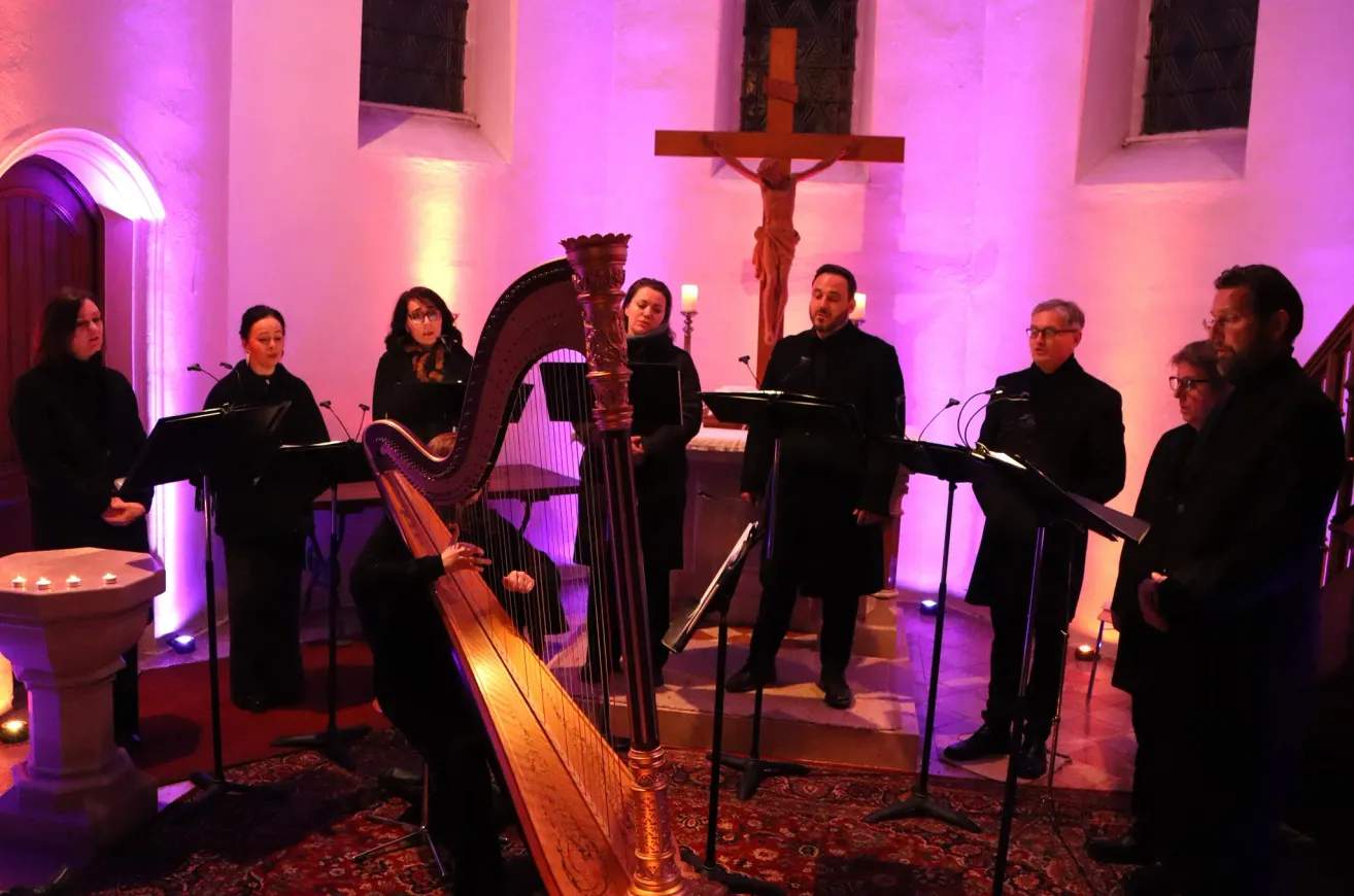 Eine Gruppe von Musikern tritt in einer Kirche auf. Eine Frau spielt die Harfe, während andere singen. Ein Kreuz und Kerzen sind im Hintergrund.