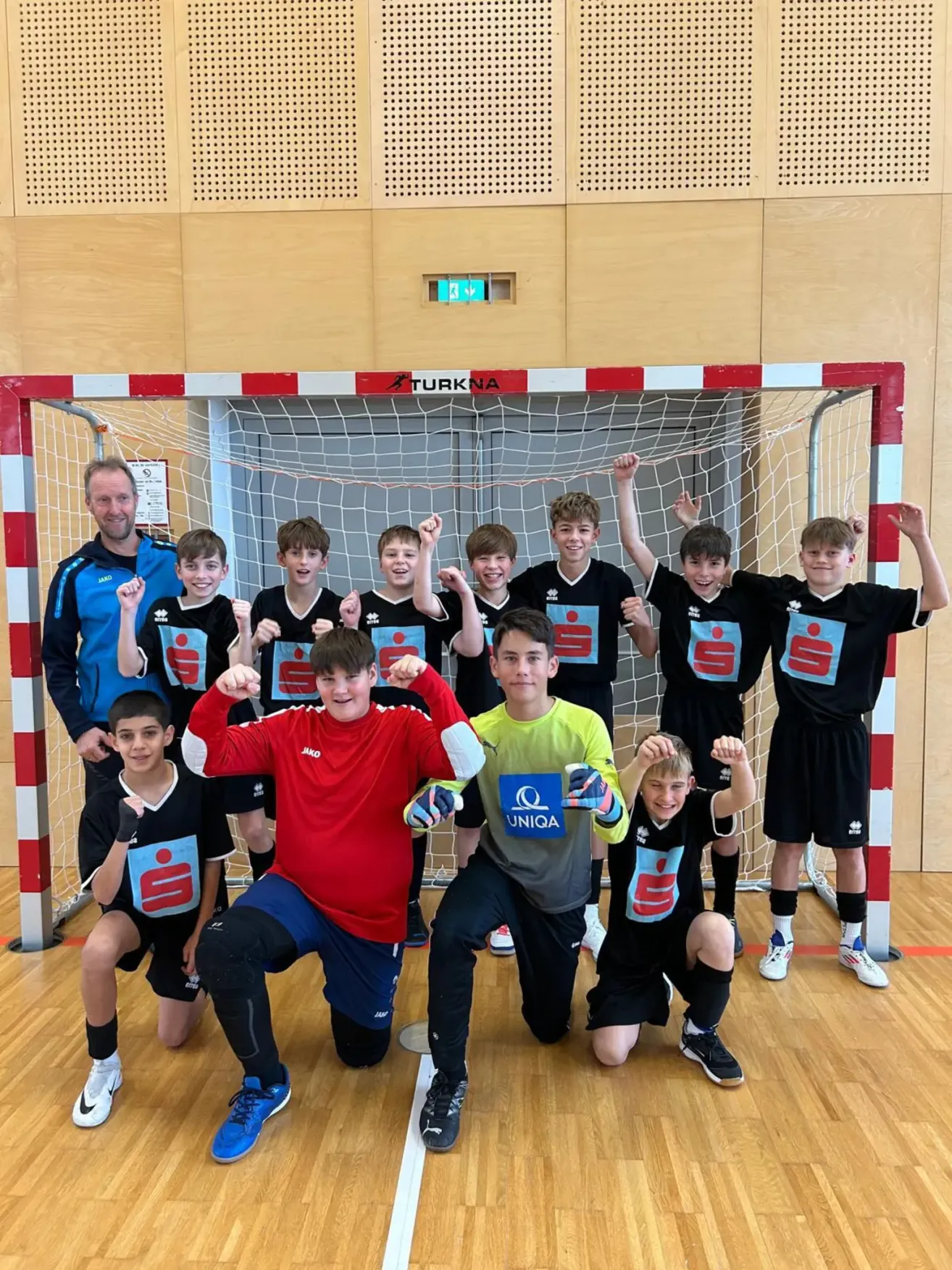 Eine Gruppe junger Jungen in Sporttrikots posiert für ein Foto vor einem Tornetz, wobei ein Mann in einem blauen Jackett und zwei Jungen in roten und gelben Trikots kniend vor ihnen stehen.