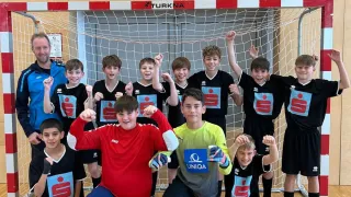 Eine Gruppe junger Jungen in Sporttrikots posiert für ein Foto vor einem Tornetz, wobei ein Mann in einem blauen Jackett und zwei Jungen in roten und gelben Trikots kniend vor ihnen stehen.