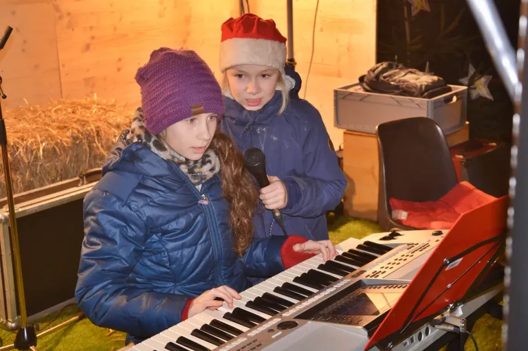 Zwei junge Mädchen spielen Klavier, eine hält ein Mikrofon, während die andere spielt. Beide tragen Winterkleidung.