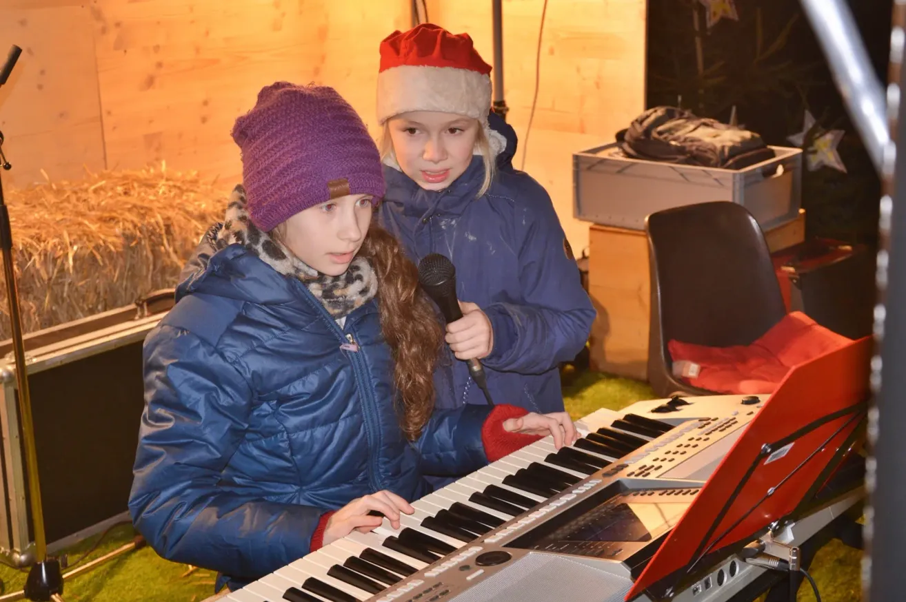 Zwei junge Mädchen spielen Klavier, eine hält ein Mikrofon, während die andere spielt. Beide tragen Winterkleidung.