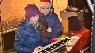 Zwei junge Mädchen spielen Klavier, eine hält ein Mikrofon, während die andere spielt. Beide tragen Winterkleidung.
