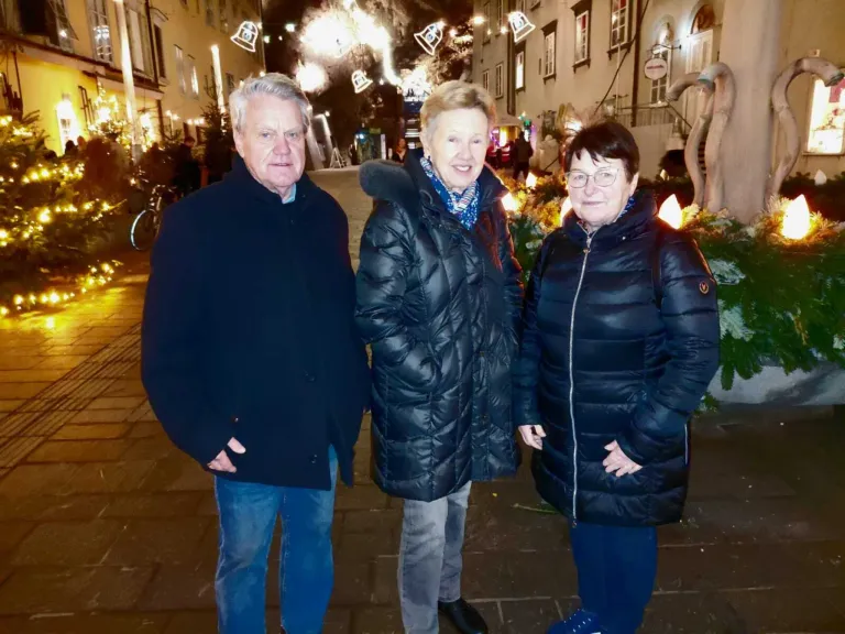 Drei Personen stehen zusammen auf einer Straße bei Nacht und lächeln für ein Foto. Sie sind warm in Wintermänteln gekleidet, und im Hintergrund sind festliche Dekorationen zu sehen.