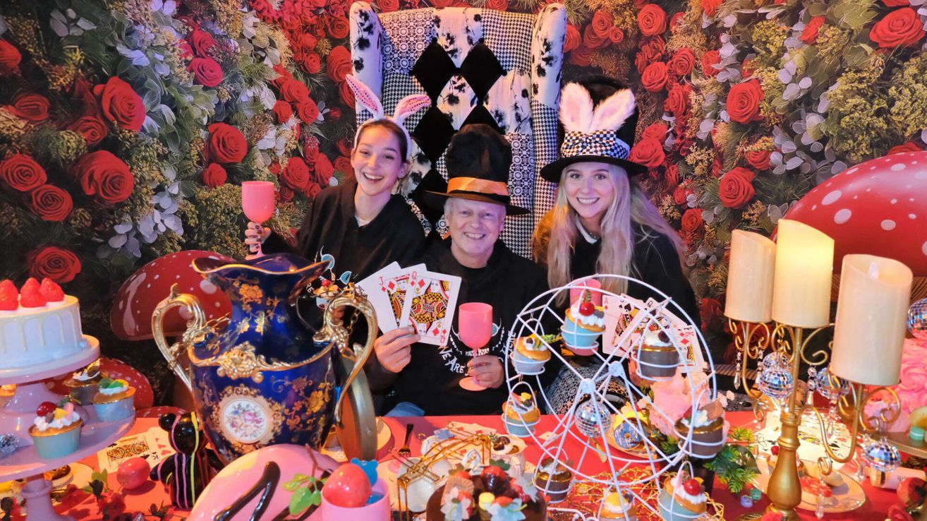 Drei Menschen sind für ein thematisches Event verkleidet. Ein Mann hält einen Becher und Spielkarten, während zwei Frauen lächeln. Ein Tisch ist mit Cupcakes, einer Vase und anderen Gegenständen dekoriert. Der Hintergrund zeigt eine Blumenwand mit einem Schachbrettstuhl.