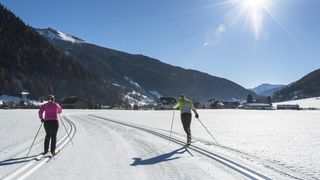 Zwei Personen betreiben Langlauf auf einem verschneiten Hang mit Bergen im Hintergrund. Sie tragen Skiausrüstung und halten Skistöcke.