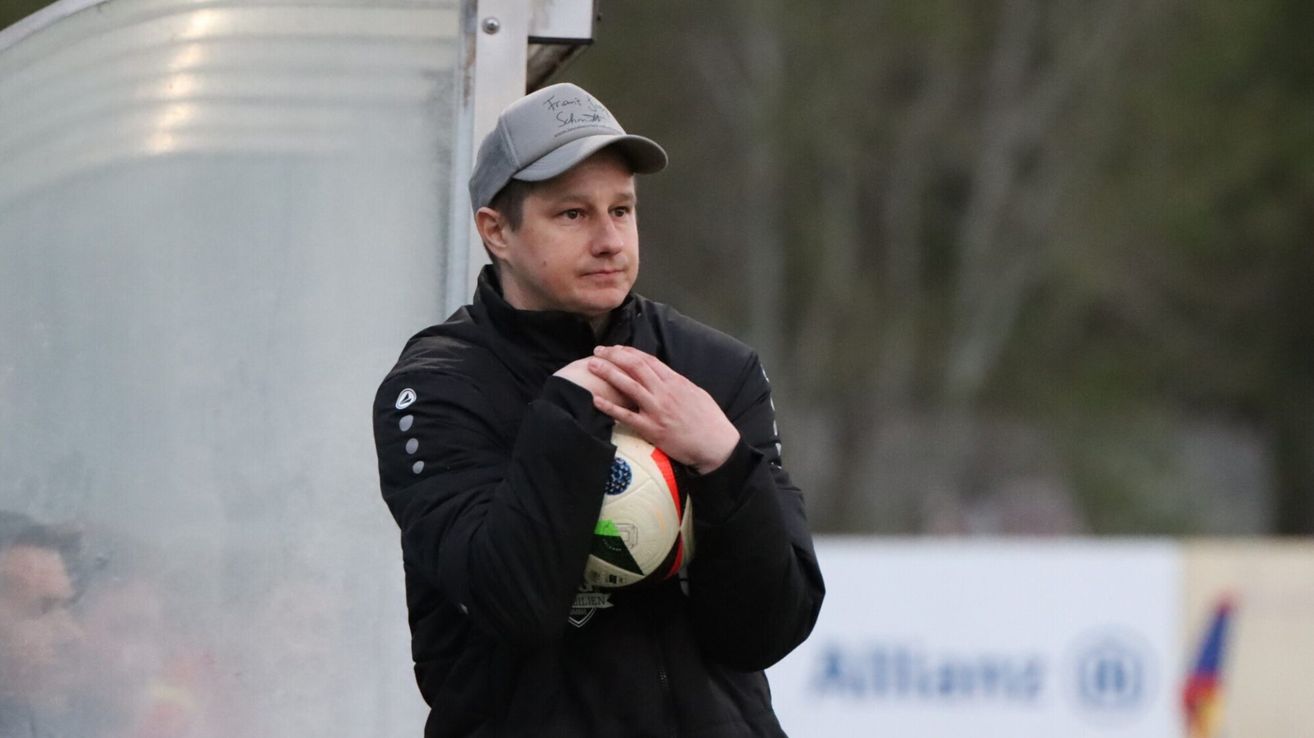 Ein Mann in einer schwarzen Jacke und einer grauen Kappe hält einen Fußball und steht vor einer weißen Wand mit verschwommenem Hintergrund.