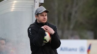 Ein Mann in einer schwarzen Jacke und einer grauen Kappe hält einen Fußball und steht vor einer weißen Wand mit verschwommenem Hintergrund.