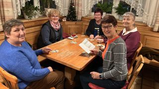 Eine Gruppe von Frauen sitzt um einen Holztisch in einem gemütlichen Raum, lächelt und hält Getränke. Ein Weihnachtsbaum und Topfpflanzen sind im Hintergrund.