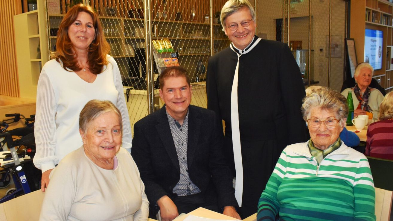 Fünf Menschen lächeln und posieren für ein Foto. Ein Mann und eine Frau sitzen, während drei andere hinter ihnen stehen. Ein Mann in einem schwarzen Gewand steht rechts, und ein Regal mit Flaschen ist hinter ihnen.