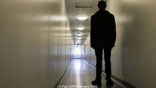 Ein Mann steht in einem Flur, den Rücken gekehrt, vor einer entfernten Ausstiegsleuchte. Der Flur ist lang und von Deckenleuchten beleuchtet.