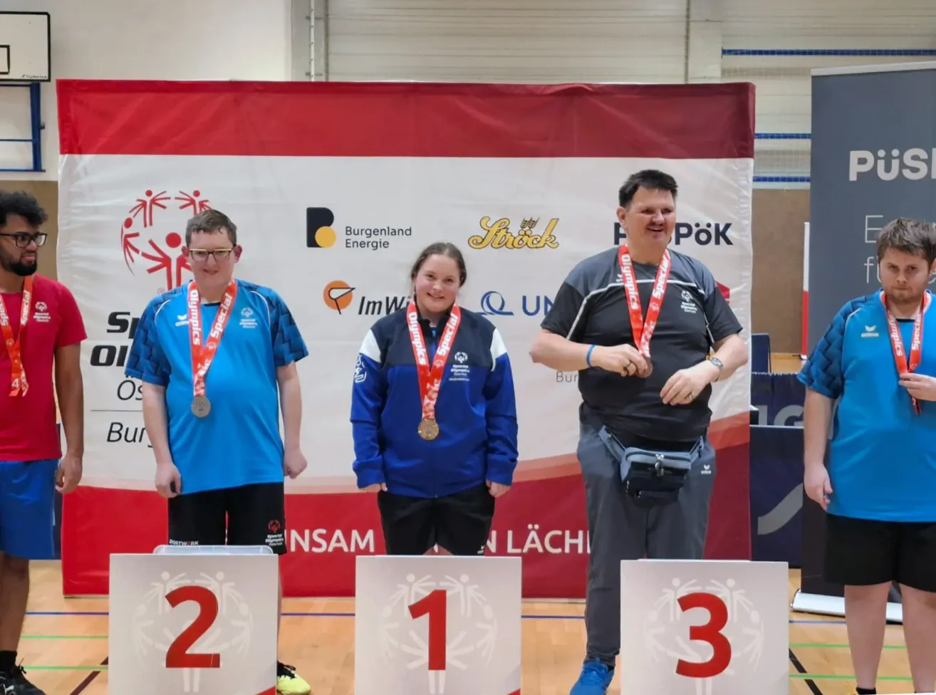 Drei Personen stehen auf einem Podest mit Medaillen, zwei in Blau und eine in Grau. Sie lächeln für ein Foto vor einem Banner.