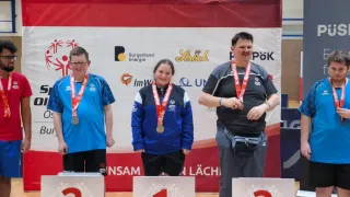 Drei Personen stehen auf einem Podest mit Medaillen, zwei in Blau und eine in Grau. Sie lächeln für ein Foto vor einem Banner.