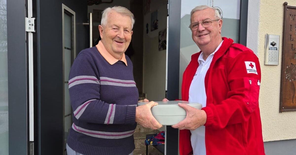 Zwei ältere Männer tauschen draußen stehend eine graue Box aus, lächeln, einer trägt einen lila Pullover und der andere eine rote Jacke.