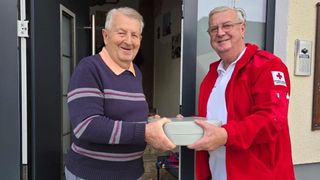 Zwei ältere Männer tauschen draußen stehend eine graue Box aus, lächeln, einer trägt einen lila Pullover und der andere eine rote Jacke.