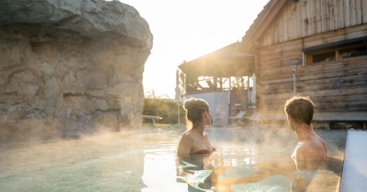 Eine Frau und ein Mann genießen ein heißes Quellenbad in der Nähe eines Holzhauses. Die Sonne geht hinter dem Gebäude unter.