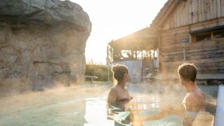 Eine Frau und ein Mann genießen ein heißes Quellenbad in der Nähe eines Holzhauses. Die Sonne geht hinter dem Gebäude unter.