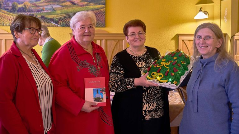 Zwei Frauen lächeln und posieren für ein Foto. Eine hält ein Buch, die andere einen dekorierten Kuchen mit Lichtern und einem festlichen Design. Der Hintergrund zeigt eine gelbe Wand mit einer Landschaftsmalerei.