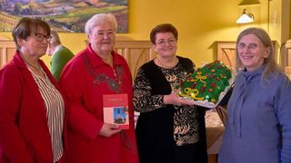 Zwei Frauen lächeln und posieren für ein Foto. Eine hält ein Buch, die andere einen dekorierten Kuchen mit Lichtern und einem festlichen Design. Der Hintergrund zeigt eine gelbe Wand mit einer Landschaftsmalerei.