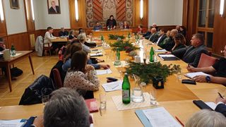 Eine Gruppe von Menschen sitzt bei einem Meeting an einem langen Tisch, mit einer Frau am Kopfende. Der Tisch ist mit Flaschen, Gläsern und Papieren besetzt. Der Raum hat eine Holzwand mit einem dekorativen Wandteppich.