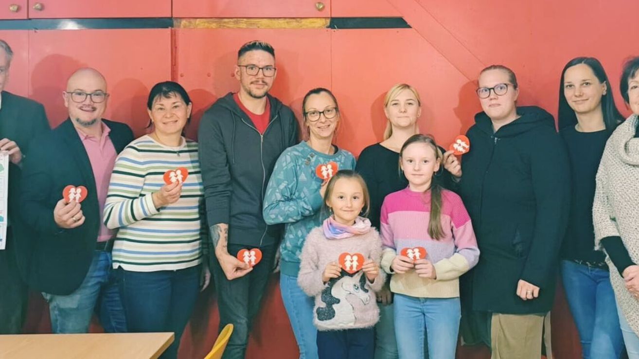 Eine Gruppe von sieben Menschen, darunter Frauen und Mädchen, stehen gemeinsam vor einer roten Tür, halten herzförmige Schilder und lächeln.