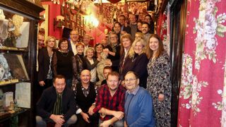 Eine Gruppe von Menschen posiert für ein Foto in einem Raum mit roten Wänden und vielen Bilderrahmen, einer Kronleuchter und einer Lampe. Einige halten eine Statue, während andere Brillen und Halsketten tragen.