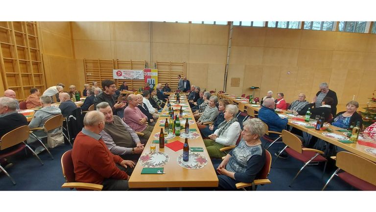 Eine Gruppe älterer Menschen sitzt an einem langen Tisch, möglicherweise bei einer Versammlung. Flaschen, Gläser und Teller stehen auf dem Tisch. Ein Banner mit Text ist an der Wand angebracht.