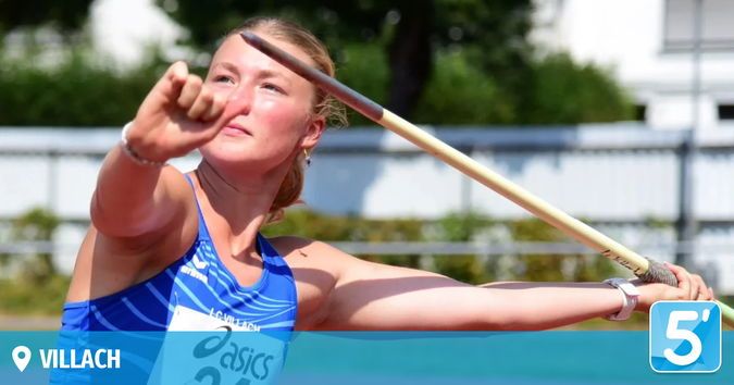 Eine Athletin wirft einen Speer, trägt ein blaues Trikot mit dem Wort 'asics' und der Nummer '23'. Sie befindet sich bei einer Sportveranstaltung im Freien.