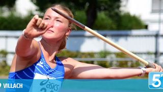 Eine Athletin wirft einen Speer, trägt ein blaues Trikot mit dem Wort 'asics' und der Nummer '23'. Sie befindet sich bei einer Sportveranstaltung im Freien.